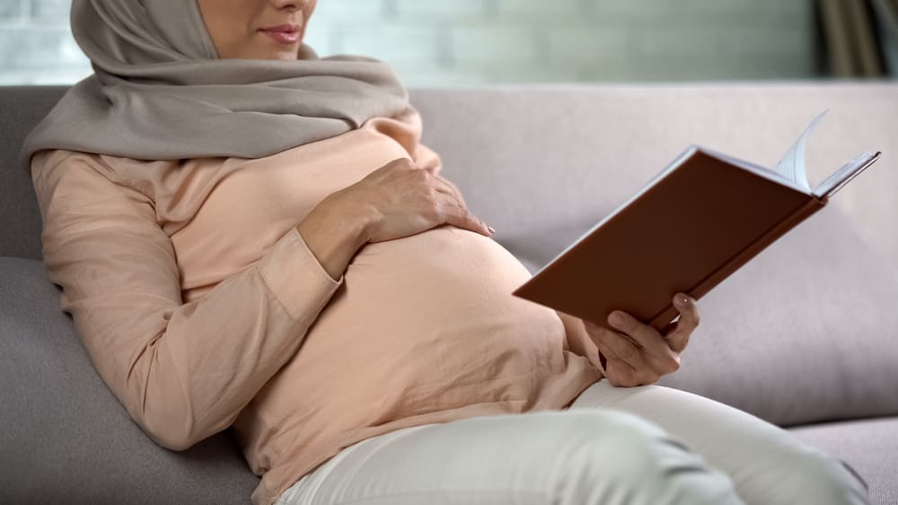 Pregnant woman in hijab stroking tummy, reading book, inner harmony, leisure