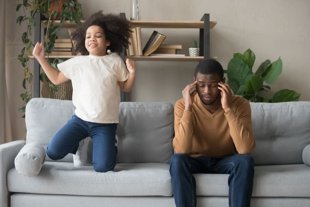 African American father having problem with noisy naughty daughter adhd girl jumping on sofa